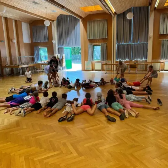 In einem Raum sitzen Kinder in einem Kreis auf dem Boden. Ein Erwachsener steht vor ihnen. Der Raum hat Holzböden, Vorhänge und Tische.