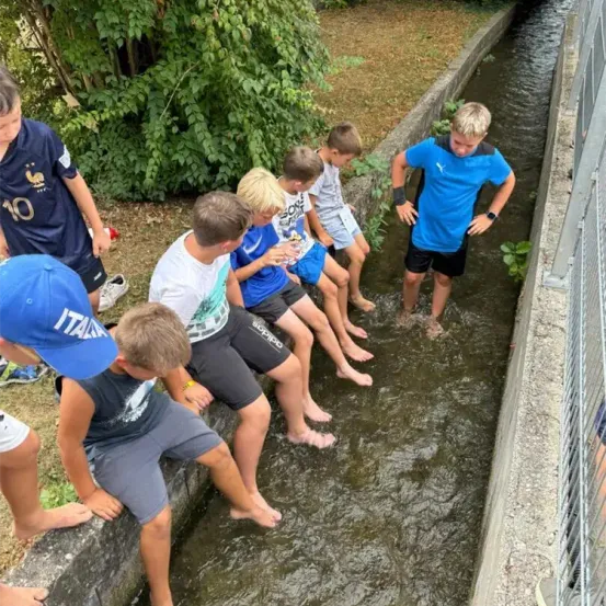 Eine Gruppe von Kindern sitzt auf einer Steinkante in einem Kanal, wobei ein Junge im Wasser steht. Sie sind alle barfuß.