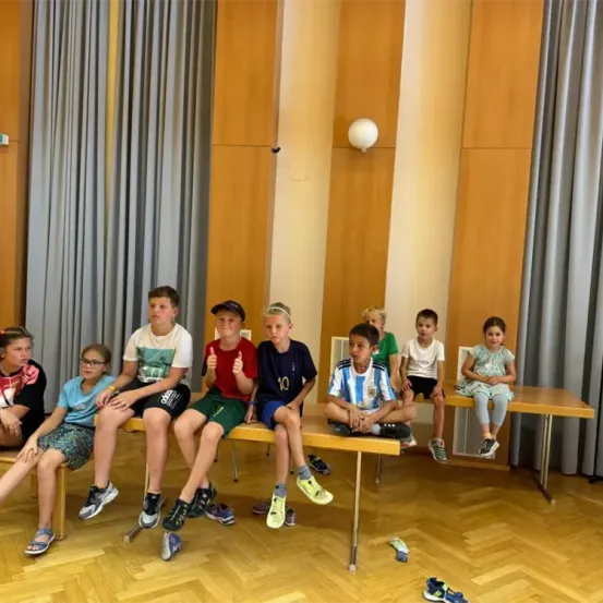 Eine Gruppe von Kindern sitzt auf Bänken in einem Raum mit Holzwänden, ein Junge trägt ein blaues Shirt mit einem Daumen hoch, und einige Schuhe liegen auf dem Boden.