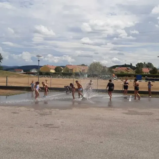 Mehrere Kinder spielen mit Wasser in einem Außenbereich. Einige planschen im Wasser, andere stehen herum. Im Hintergrund befinden sich Häuser, und der Himmel ist bewölkt.