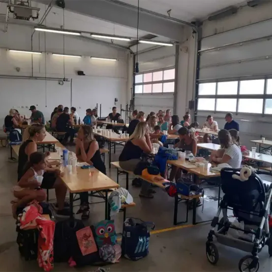 Eine Gruppe von Menschen sitzt um Tische in einem großen, offenen Raum. Einige tragen Freizeitkleidung, und einige haben Taschen und Kinderwagen in der Nähe. Der Raum ist gut beleuchtet mit Deckenleuchten und großen Fenstern.