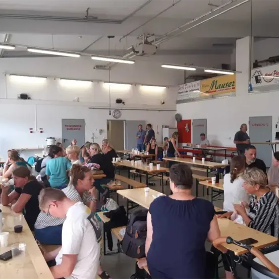 Ein überfüllter Innenbereich mit Esstischen, an denen Menschen sitzen, die Gespräche führen, mit Tassen und Tellern auf den Tischen und einer Uhr und einem Banner an der Wand.