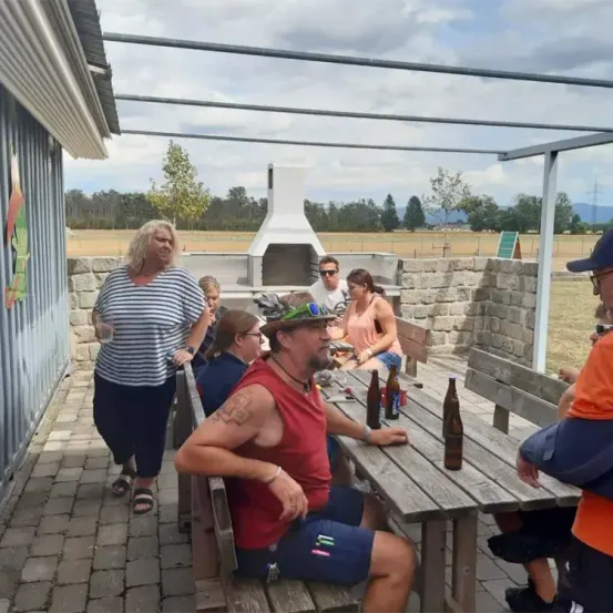 Eine Gruppe von Menschen sitzt um einen Tisch auf einer Terrasse, einige tragen Hüte und Sonnenbrillen. Eine Frau steht in der Nähe und hält ein Glas. Im Hintergrund befindet sich eine Steinmauer, ein Kamin und ein Grasfeld mit Bäumen.