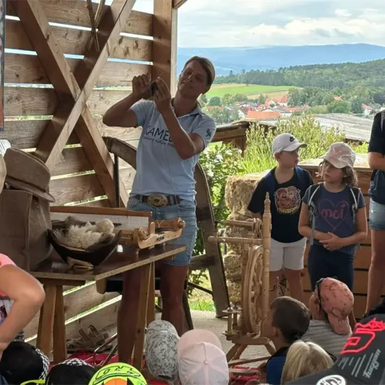 Eine Frau in einem T-Shirt demonstriert den Gebrauch eines Spinnrades einer Gruppe von Kindern in einer rustikalen Umgebung mit einer landschaftlichen Kulisse.