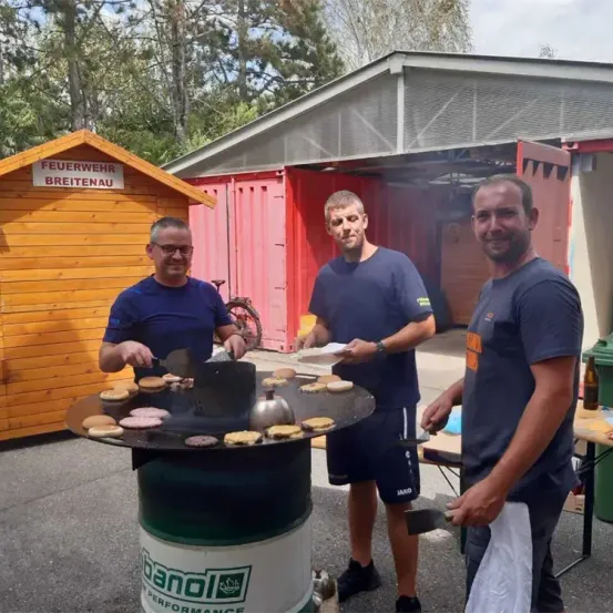 Drei Männer kochen Essen auf einem Grill im Freien. Ein Mann hält eine Pfanne, ein anderer einen Tablett und der dritte ein Tuch. Sie stehen vor einem Feuerwehrgebäude.