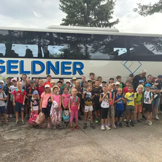 Eine Gruppe von Kindern mit Rucksäcken steht vor einem Bus mit der Aufschrift Geldner. Der Bus ist auf einem Parkplatz geparkt.