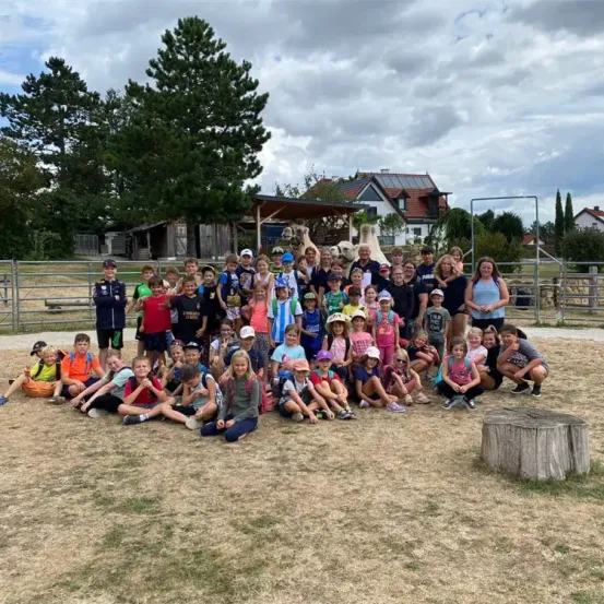 Eine Gruppe von Kindern und Erwachsenen versammelt sich auf einem Feld, einige sitzen und andere stehen, mit einigen Kamelen hinter einem Zaun. Bäume und Häuser sind im Hintergrund zu sehen.