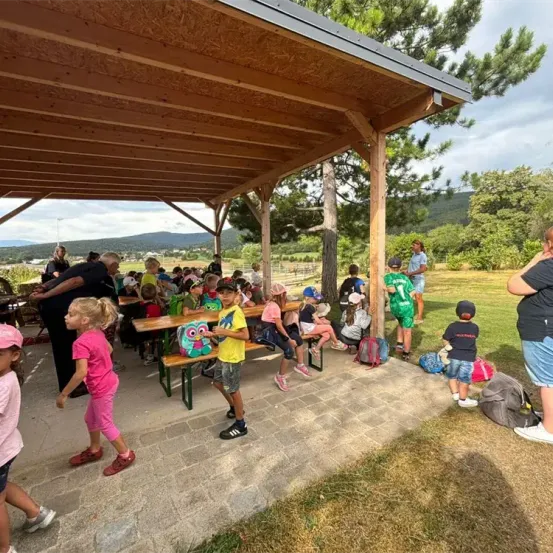Eine Gruppe von Kindern und Erwachsenen sitzt unter einem Pavillon in einem Außenbereich. Einige von ihnen tragen Hüte und Turnschuhe. Eine Frau steht hinter ihnen.