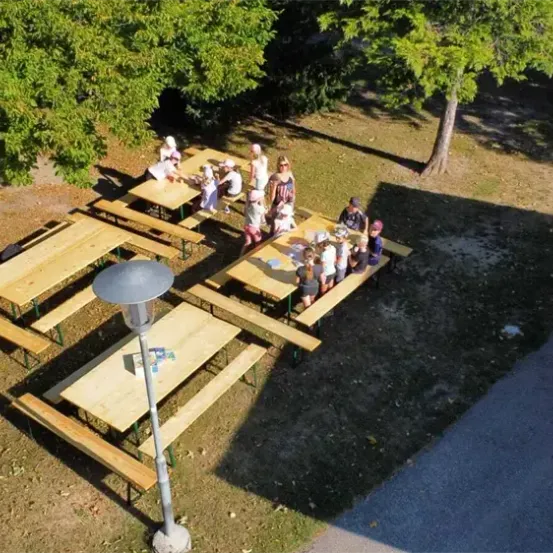 Eine Gruppe von Menschen sitzt um einige Holztische in einem Außenbereich, möglicherweise einem Park. Einige tragen Hüte und Sonnenbrillen. Der Bereich ist von Bäumen und Gras umgeben.