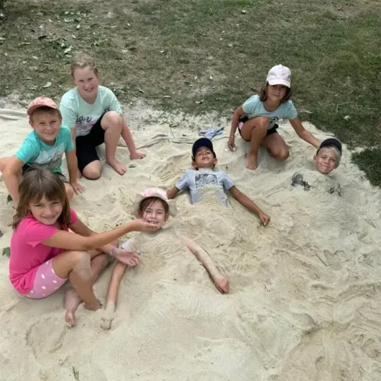 Eine Gruppe von Kindern spielt im Sand, einige sitzen und einige liegen, alle lächeln. Gras ist hinter ihnen sichtbar.
