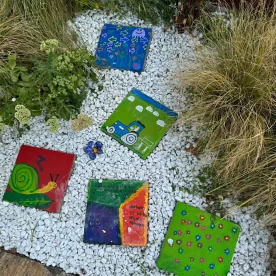 Ein Garten mit sechs bemalten Fliesen auf weißen Kieselsteinen, mit verschiedenen Designs wie eine Schnecke, ein Lastwagen, Fische und Blumen.