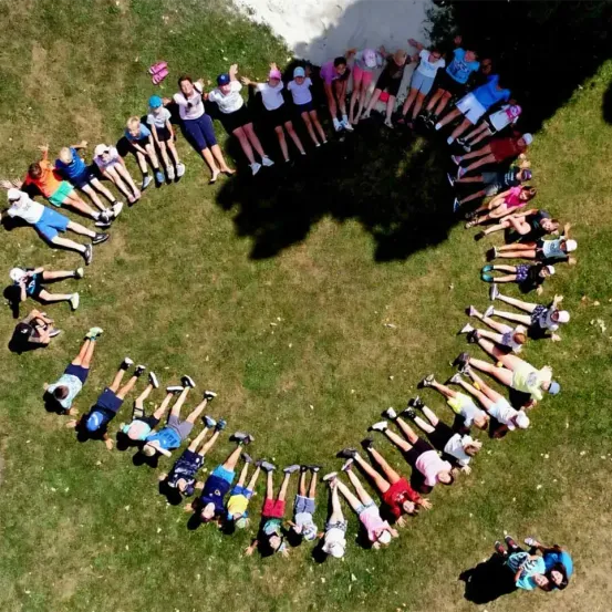 Eine Gruppe von Kindern und Erwachsenen bildet einen Kreis auf einem Rasenfeld. Sie tragen lässige Sommerkleidung und einige haben Mützen auf. Das Bild ist von oben aufgenommen.