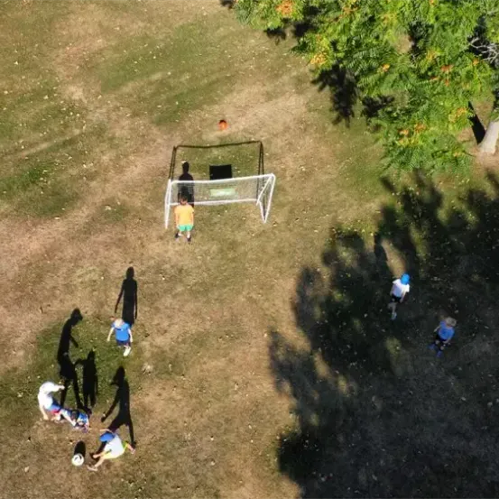 Eine Draufsicht auf ein saftiges Feld zeigt eine Gruppe junger Spieler, die Fußball trainieren. Sie sind um ein Tor herum positioniert, und einige treten einen Ball.