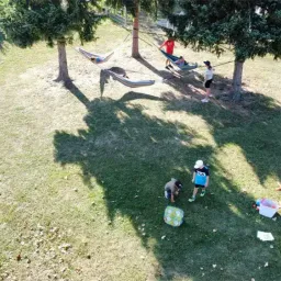 Eine Vogelperspektive eines Grasfelds zeigt Menschen in Hängematten und andere, die herumstehen, mit Spielzeug und einem Eimer in der Nähe.