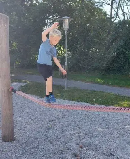 Ein junger Junge läuft auf einem Seil auf einem Spielplatz. Bäume und eine Straßenlaterne sind im Hintergrund zu sehen.