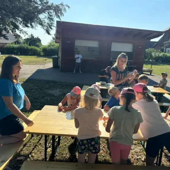 Eine Gruppe von Kindern und Erwachsenen sitzt um Tische in einer Outdoor-Umgebung. Eine Frau vor ihnen scheint eine Aktivität zu leiten. Das Gebiet ist von Bäumen und einem kleinen Gebäude umgeben.