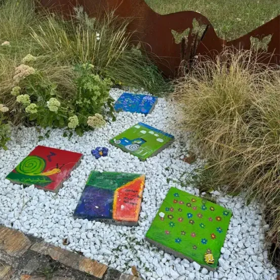 Ein Garten mit einem Rand aus rostigen Eisenblechen und verschiedenen farbigen Fliesen mit Zeichnungen, auf weißen Kieseln platziert.