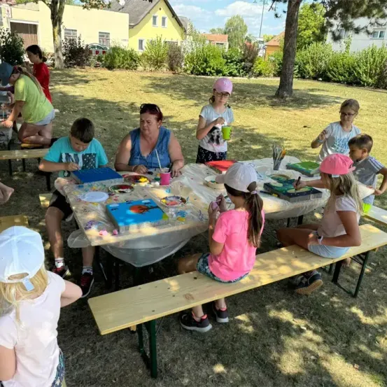 Mehrere Kinder und Erwachsene sitzen um einen Tisch im Freien und malen. Sie sitzen auf Bänken, eine Person steht. Eine Frau hält einen Pinsel.