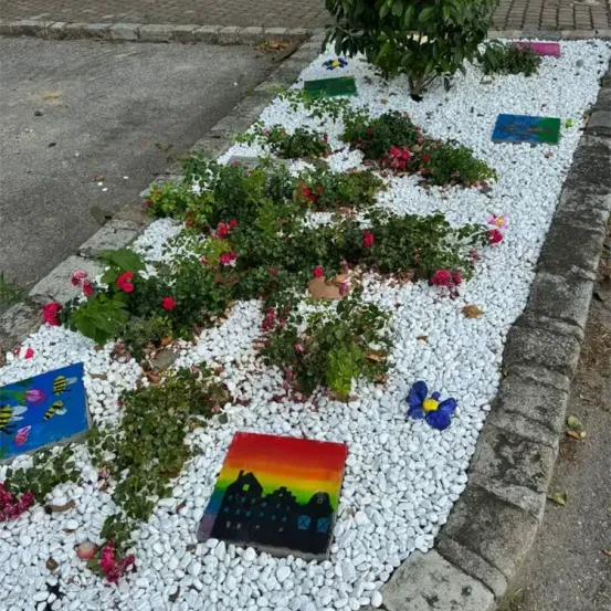Ein Gartenbeet gefüllt mit weißen Kieselsteinen und verschiedenen Pflanzen, darunter rote Blumen, grüne Blätter und eine bunte Malerei auf einem Stein. Das Beet ist von Ziegelsteinen gesäumt und hat einen kleinen Baum an der Seite.