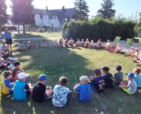 Eine Gruppe von Kindern und Erwachsenen sitzt im Kreis auf einem Grasfeld, einige tragen Mützen. Eine Frau steht in der Nähe eines weißen Hauses mit Schornsteinen.
