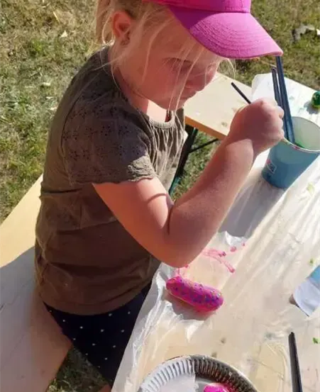 Ein kleines Mädchen malt mit verschiedenen Farben auf einem Teller im Freien. Sie trägt eine rosa Mütze und ein braunes Shirt. Auf dem Tisch befinden sich ein Becher und Pinsel. Sie sitzt auf einer Bank.