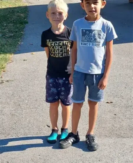 Zwei junge Jungen stehen auf einem gepflasterten Weg, einer trägt ein schwarzes T-Shirt mit grafischem Design und der andere einen blauen Hut und ein blaues T-Shirt. Sie tragen beide Turnschuhe.