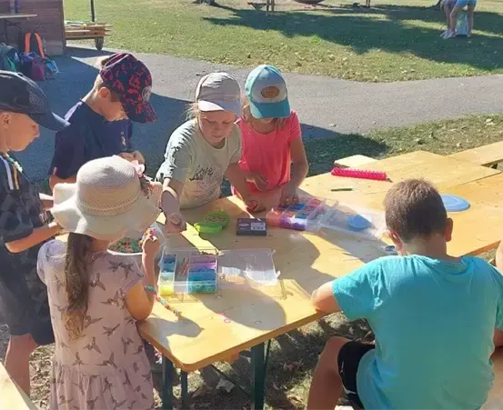 Eine Gruppe von Kindern sitzt um einen Tisch in einem Park und bastelt mit verschiedenen Materialien. Eine Frau beaufsichtigt sie.