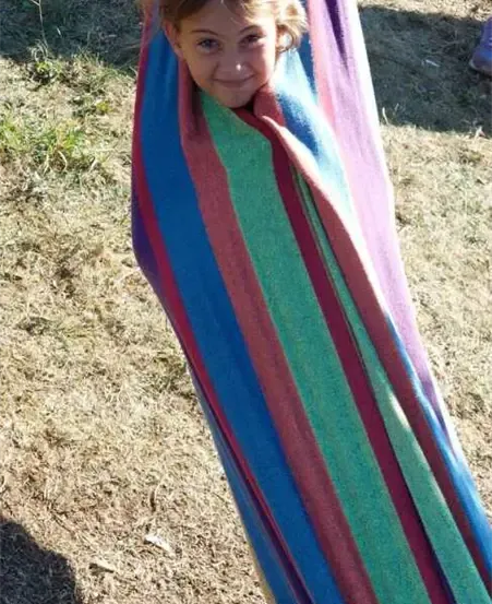 Ein junges Mädchen mit blondem Haar liegt in einem farbenfrohen Hängesessel an einem sonnigen Tag. Der Hängesessel ist an einem Baum aufgehängt, und das Mädchen lächelt.