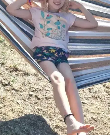Ein kleines Mädchen liegt auf einer bunten Hängematte und lächelt. Sie trägt ein T-Shirt mit Blumenmuster und Shorts. Der Boden ist mit trockenem Gras bedeckt.