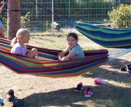 Drei junge Mädchen entspannen sich in Hängematten. Zwei sitzen, während die andere in der Nähe steht und eine Schaukel hält. Das Gebiet ist von einem Zaun umgeben, mit Schuhen und einem Helm verstreut.