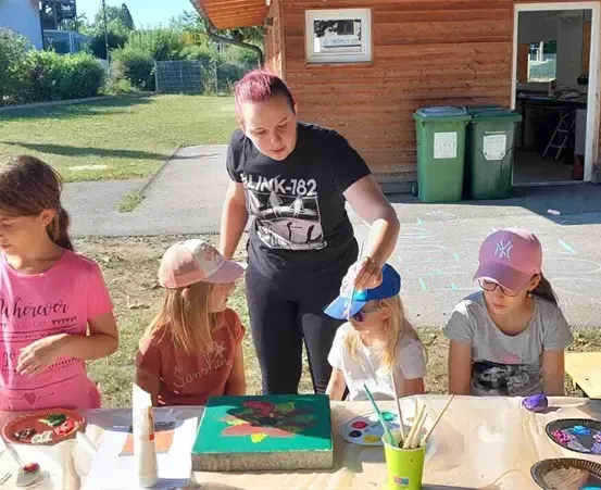 Eine Frau unterrichtet Kinder im Malen im Freien. Vier Kinder sitzen an einem Tisch, und ein Holzhaus ist im Hintergrund.