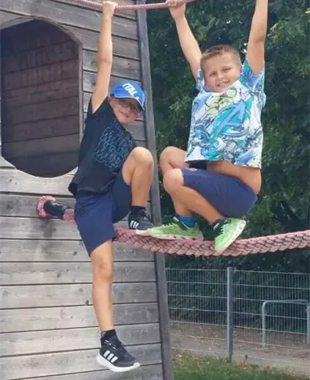 Zwei Jungen hängen an einem Seilschwinger an einer Holzkonstruktion. Ein Junge trägt einen Hut und Turnschuhe, der andere ein T-Shirt mit grafischem Design. Sie tragen beide Shorts.