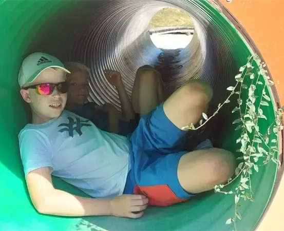 Ein Junge liegt in einem grünen Röhrenrutsche, trägt eine grüne Mütze und Sonnenbrille. Ein weiterer Junge sitzt neben ihm.