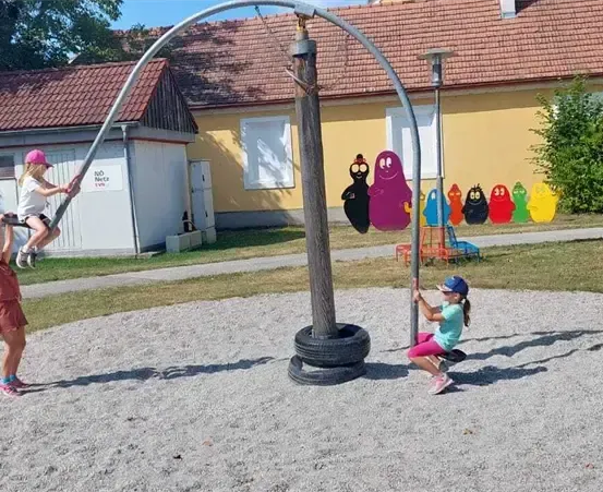 Drei Kinder spielen auf einem Spielplatz mit einer Reifenschaukel und bunten Charakteren vor einem gelben Gebäude.