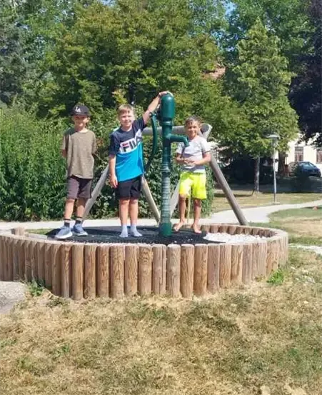 Drei Jungen stehen um eine große grüne Pumpe in einem Park mit Gras und Bäumen.