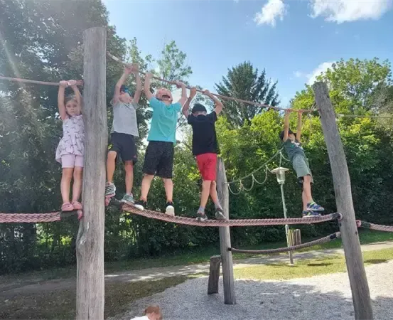 Fünf Kinder hängen an einer horizontalen Stange in einem Freiluft-Spielplatz mit grünem Rasen und Bäumen im Hintergrund.