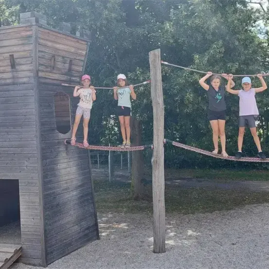 Vier Mädchen balancieren auf Seilen in einem Spielplatz. Eines hält sich an einem Holzturm fest. Sie sind draußen.