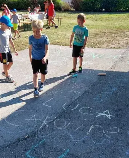 Kinder stehen um eine Kreidezeichnung auf dem Boden herum, die 'USA Around Team' besagt. Im Hintergrund sind mehrere Häuser und ein Picknick-Setup zu sehen.