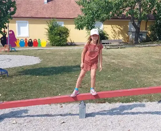 Drei junge Mädchen spielen auf einer Wippe in einem Park, mit einem Haus und farbenfrohen Charakteren, die an der Wand im Hintergrund gemalt sind.