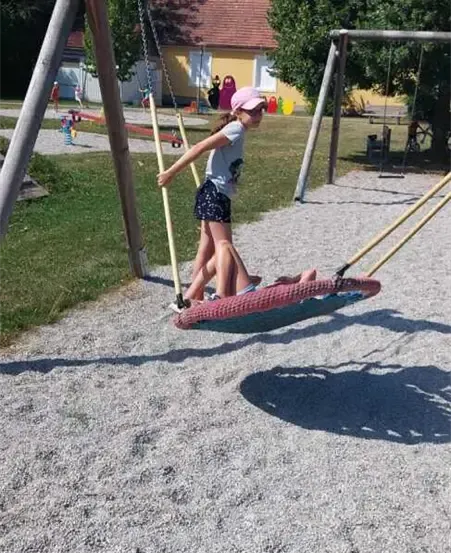 Zwei Kinder spielen auf einer Schaukel in einem Park, eine weitere Schaukel befindet sich in der Nähe. Dahinter steht ein Haus mit rotem Dach und eine Bank unter einem Baum.