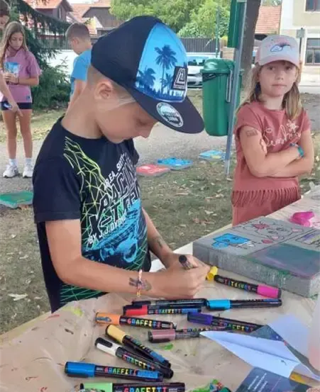 Ein junger Junge malt auf einem Tisch mit verschiedenen Markern und Pinseln. Neben ihm beobachtet ein Mädchen. Im Hintergrund befinden sich mehrere Kinder und ein Mülleimer.
