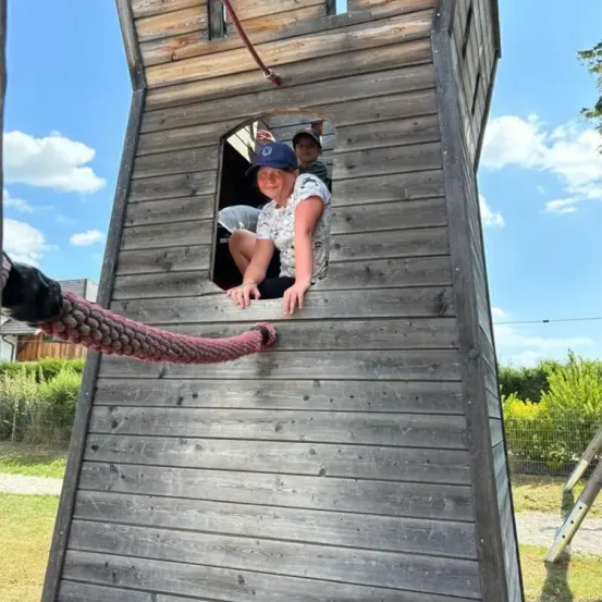 Zwei Personen befinden sich in einem Holzspielhaus. Eine Frau mit einer Mütze schaut aus dem Fenster. Ein Junge mit einer Kappe sitzt auf der Fensterbank.