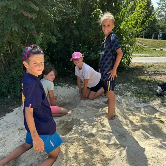 Vier Kinder spielen in einem sandigen Bereich, eines kniet und lächelt, ein anderes steht mit den Händen auf den Hüften und zwei andere sitzen. Dahinter sind Grünflächen und ein Weg sichtbar.