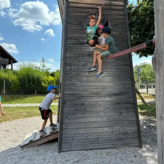 Vier Kinder spielen auf einer Holzstruktur. Drei sitzen darauf, während eines hinaufklettert. Sie tragen Mützen und Turnschuhe. Die Umgebung ist draußen, mit Gras, Büschen und Bäumen sichtbar.