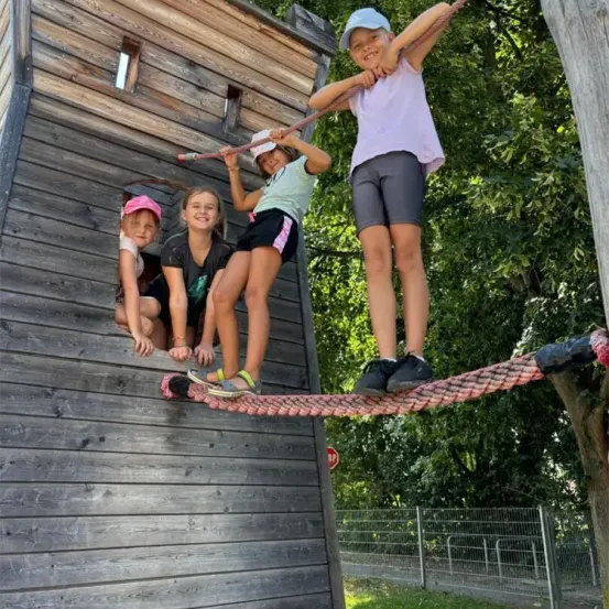 Vier Kinder klettern eine an einem Holzgebäude befestigte Seilleiter hinauf, während ein Mädchen mit einer rosa Mütze aus einem Fenster zuschaut.