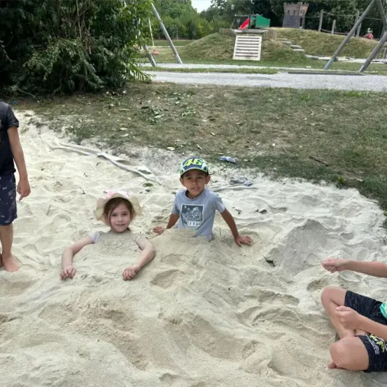Vier Kinder spielen in einem Sandkasten, wobei zwei im Sand vergraben sind und zwei stehen. Dahinter ist ein grasbewachsener Hügel mit einer Holzspielstruktur zu sehen.