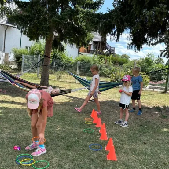 Kinder spielen auf einem Grasfeld mit bunten Hula-Hoops und Kegel. Eine Frau beugt sich nach unten, um einen Hula-Hoop aufzuheben. Ein Hängematten zwischen zwei Bäumen steht hinter ihnen.