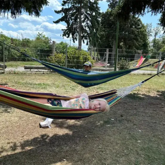 Zwei Kinder liegen in einer Hängematte in einem Park mit Zaun und Bäumen. Ein Kind lächelt.