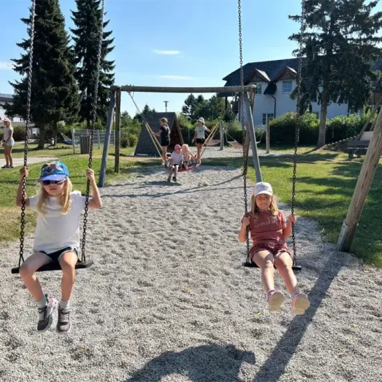 Zwei Mädchen schwingen auf Schaukeln in einem Spielplatz, eines trägt ein weißes Shirt und das andere ein rotes. Dahinter spielen andere Kinder auf Schaukeln.