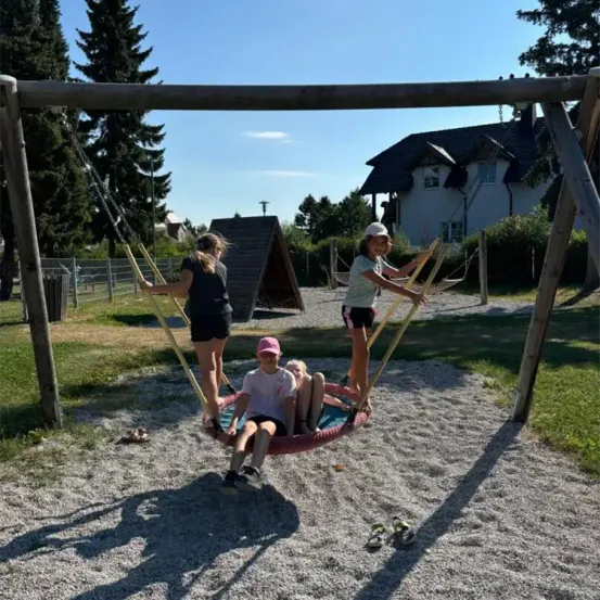Vier Kinder spielen auf einer Reifenschaukel in einem Park, zwei schwingen, während zwei die Seile halten. Dahinter steht ein Haus mit Schornstein und einer Hütte. Bäume und Büsche sind im Hintergrund zu sehen.